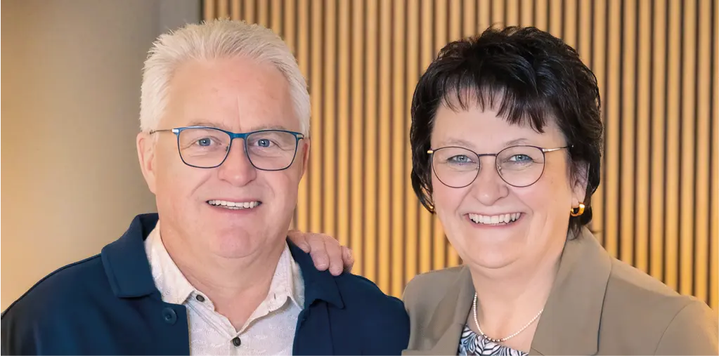 Zwei Personen stehen nebeneinander vor einer hellen Lamellenwand, beide tragen Brillen und helle Oberteile, eine Person mit dunklem Blazer, eine mit dunkler Jacke.